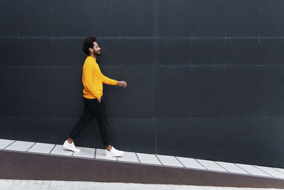 Smiling man walking with hand in pocket on footpath by black wall