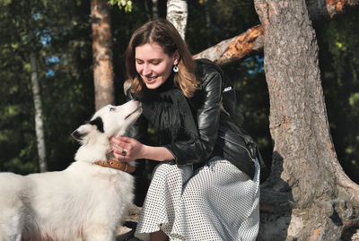 Front view of young smiling woman stroking her dog whle sitting under the tree in a forest.