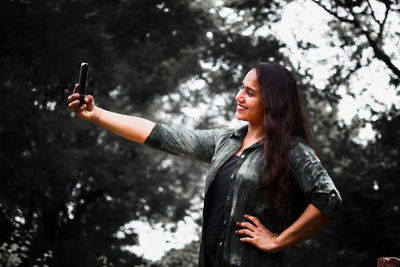 Smiling young woman taking selfie with phone while standing against tree