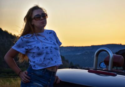 Beautiful woman standing by car against sky during sunset