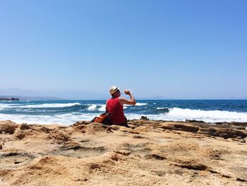 Rear view of man drinking water while sitting at shore of beach against sky