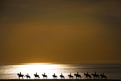 Silhouette people riding horses on sea shore against sky during sunset