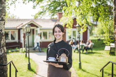 Portrait of confident young female waitress smiling while holding serving tray against outdoor cafe