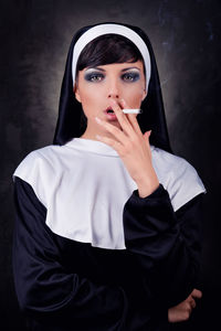 Portrait of nun smoking while standing against black background