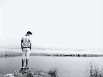 Full length of man standing on rock at lakeshore against clear sky