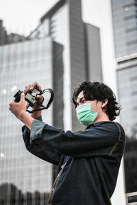 Man wearing mask holding camera while standing against building in city