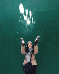 Young woman throwing snow while standing against green wall