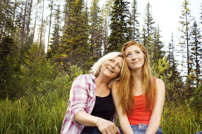 Thoughtful smiling mother and daughter sitting against trees at forest