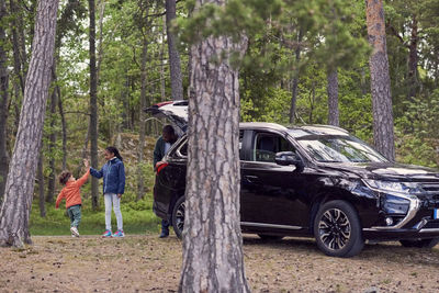 Father looking at siblings giving high-five while standing by car amidst trees