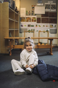 Portrait of smiling girl tying shoelace while sitting on floor in classroom at kindergarten