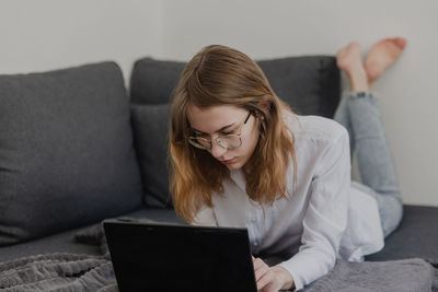 Attractive young woman working from home - female entrepreneur sitting on sofa with laptop computer and checking cell phone from comfort of home