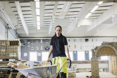 Female carpentry student pushing wooden planks in wheelbarrow at site