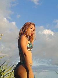 Low angle view of young woman in bikini standing against cloudy sky