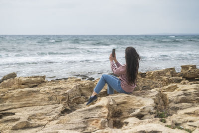 Rear view of woman sitting on rock at beach against sky