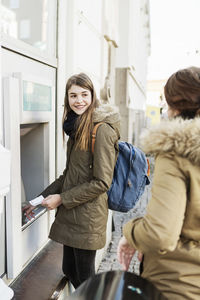 Happy young woman looking at female friend while standing at cash machine