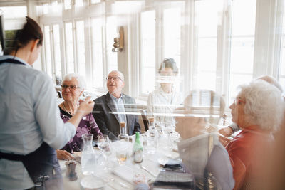 Female owner talking with senior customer sitting with friends seen through glass window