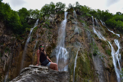 Low angle portrait of mid adult woman wearing sunglasses sitting on rock against waterfall