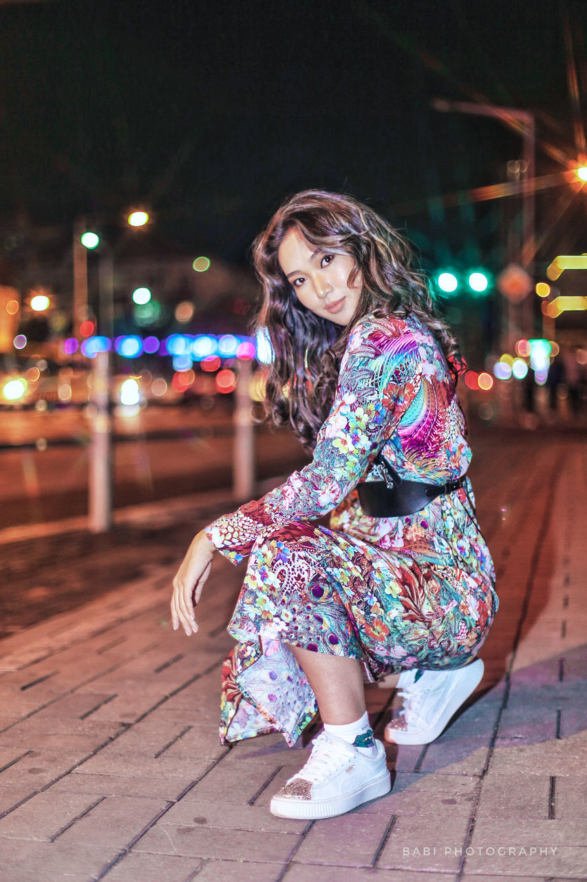 one person, full length, night, looking at camera, lifestyles, street, real people, young adult, focus on foreground, illuminated, city, women, leisure activity, portrait, young women, casual clothing, architecture, adult, outdoors, beautiful woman, hairstyle, floral pattern