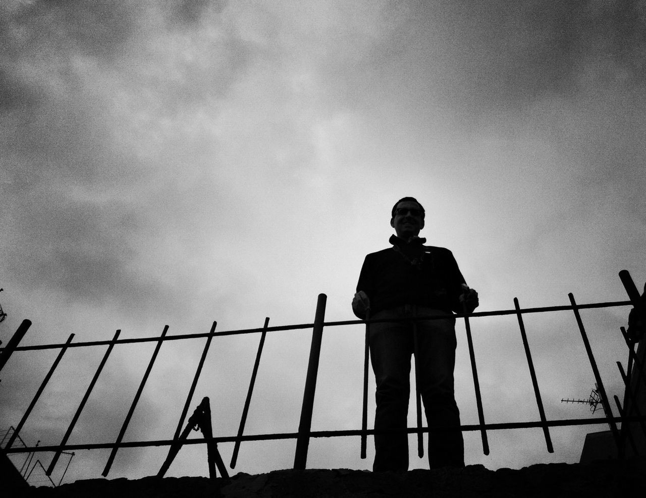 sky, low angle view, silhouette, fence, railing, protection, standing, lifestyles, safety, men, leisure activity, metal, security, cloud - sky, chainlink fence, full length, cloud, built structure