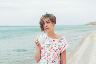 Thoughtful woman holding coffee cup while standing at beach against sky