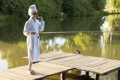 Caucasian woman in white robe and towel is fishing holding spinning rod on pier of lake early in the