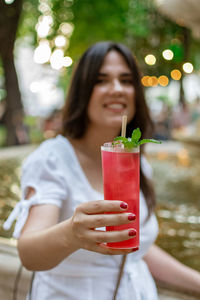 Beautiful young woman drinking strawberry mojito cocktail in park in city