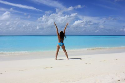 Full length rear view of woman with arms raised standing at beach against sky