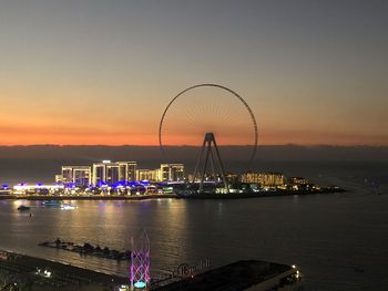 Illuminated ferris wheel by sea against sky during sunset