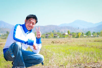 Portrait of smiling man showing peace sign while crouching in field