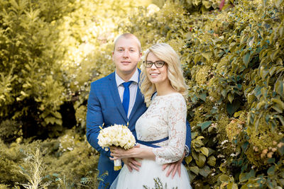 Portrait of smiling bride and groom standing against plants at park