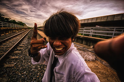 Portrait of man gesturing on railroad track against cloudy sky