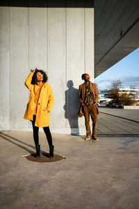 Full length of black woman with afro curly hair wearing vivid coat standing with hands in pocket looking away on street with cool guy on background