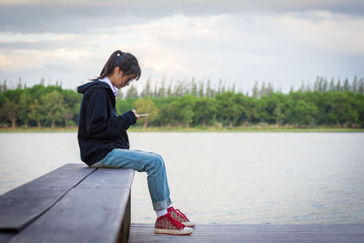 Side view of woman sitting by lake against sky