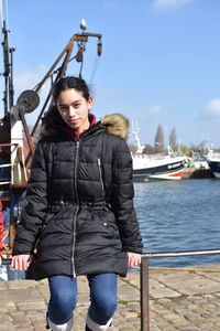 Portrait of girl wearing warm clothing while sitting on railing at harbor