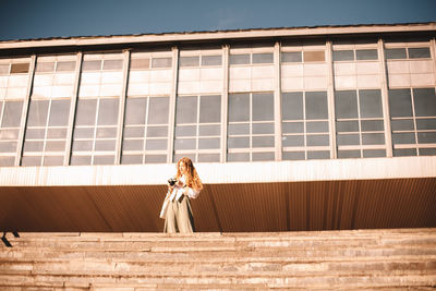 Young woman holding camera while standing in city during summer