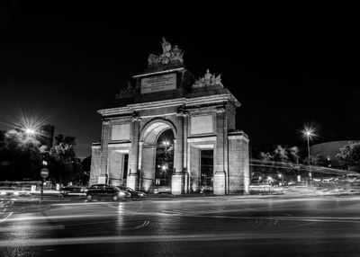 Light trails on illuminated street by puerta de toledo against sky at night