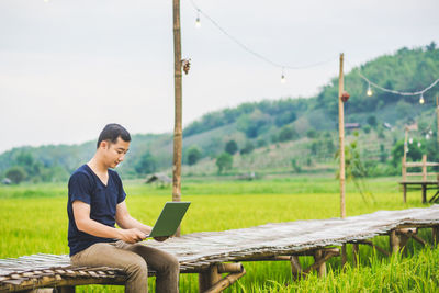 Man using laptop while sitting on boardwalk at rice paddy