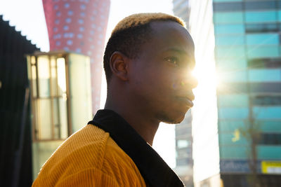 From below side view of serious african american male with dyed hair looking away while standing near modern multistory building with glass wall in city