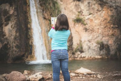 Rear view of girl photographing of waterfall through mobile phone