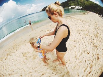 Fish-eye lens view of mother assisting daughter wearing swimming goggles at beach