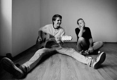 Smiling young man playing guitar by friend on hardwood floor at home