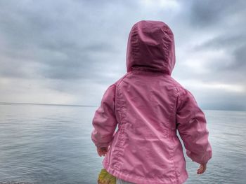 Rear view of baby girl in pink raincoat standing by lake geneva against cloudy sky