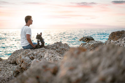 Man and french bulldog dog sitting on the beach at sunset