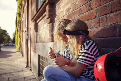 Girls using mobile phone while sitting against brick wall on sunny day
