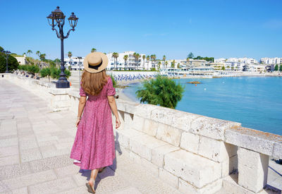 Back view of young woman with hat and dress walks towards the promenade of otranto, apulia, italy