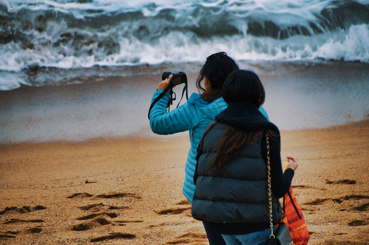 REAR VIEW OF WOMAN PHOTOGRAPHING SEA WATER FROM BEACH