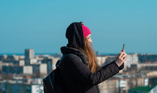 Confident woman in pink hat with smartphone in hand calling friend walking city background blue sky