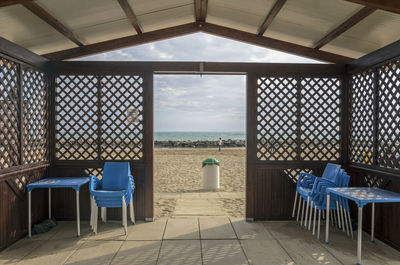 Empty chairs and tables by window at beach against sky