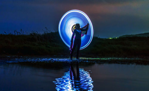 Woman spinning illuminated lightning equipment while standing by lake against sky at night