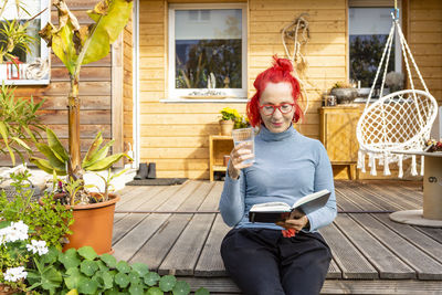 Portrait of smiling senior woman with red dyed hair sitting on terrace in front of her house reading a book